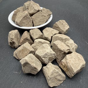 May include: A collection of light beige, irregularly shaped clay chunks. Some pieces are piled inside a white bowl, while others are scattered around the base of the bowl. Each piece features a rough, textured surface with visible natural edges.