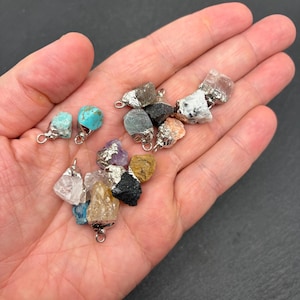 Pu&ograve; includere: Una collezione di piccoli ciondoli in pietre grezze con forme irregolari e non levigate, in vari colori tra cui turchese, quarzo trasparente, nero, giallo e viola, ciascuno dotato di un gancio in metallo argentato per essere appeso.