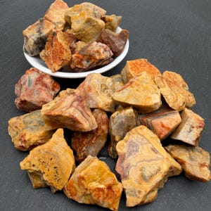 May include: A collection of raw, unpolished agate stones featuring banded patterns in shades of orange, yellow, cream, and brown. Some stones are contained within a white bowl, while others are scattered around the base of the bowl.
