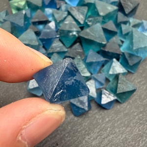 May include: A collection of small, translucent blue fluorite crystals shaped into octahedrons. Each crystal features a geometric, eight-sided form with smooth, angled facets that catch the light, displaying various shades of deep and light blue.