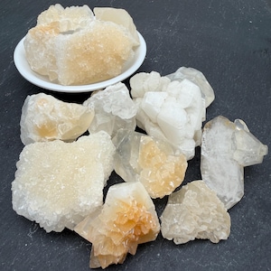 May include: A collection of raw, crystalline mineral specimens in shades of white, cream, and pale yellow. Several clusters feature jagged, translucent surfaces and geometric formations, with one group contained within a small, round white dish.