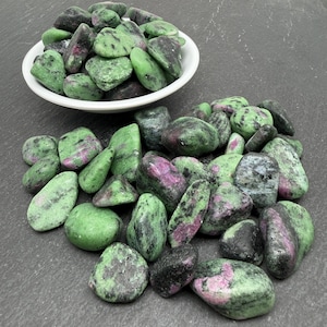 Può includere: Una collezione di pietre di zoisite levigate con inclusioni di rubino, caratterizzate da un mix di colori verde vibrante e magenta-rosa intenso. Alcune pietre sono contenute in una piccola ciotola bianca, mentre altre sono sparse alla base.