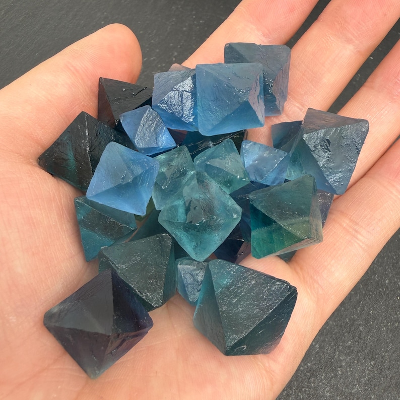 May include: A collection of small, translucent, octahedral fluorite crystals in various shades of blue, ranging from light sky blue to deep navy and teal. The crystals have sharp, geometric edges and smooth, glassy surfaces with natural inclusions.