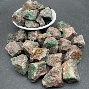 May include: A collection of raw, unpolished mineral stones featuring a mix of earthy red, pink, and vibrant green hues. Some stones are contained within a small white bowl, while others are scattered around the base of the bowl on a dark surface.