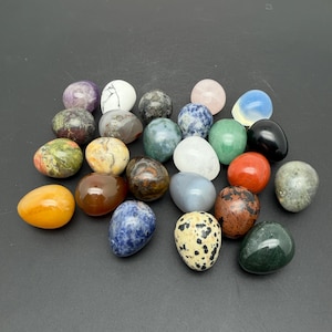 May include: A collection of twenty-two smooth, egg-shaped polished stones in various colours and patterns, including shades of white, black, green, blue, orange, brown, and grey, arranged in a cluster on a solid dark grey surface.