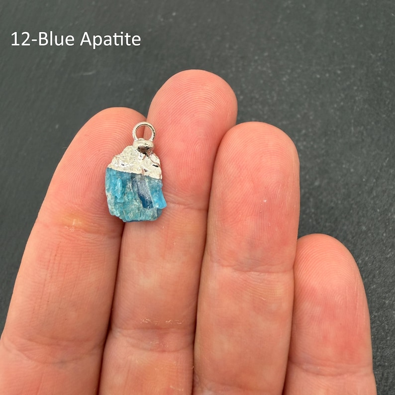Pu&ograve; includere: Un ciondolo in apatite blu grezza caratterizzato da una consistenza cristallina ruvida in tonalit&agrave; blu brillante, sormontato da un gancio in metallo color argento. Il testo 12-Blue Apatite &egrave; visibile nell'angolo in alto a sinistra.