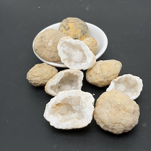 Puede incluir: Una colección de rocas geodas redondas de color tostado con exteriores rugosos y texturizados. Varias de las piedras están abiertas para revelar centros huecos llenos de cristales de cuarzo blanco translúcidos y brillantes.