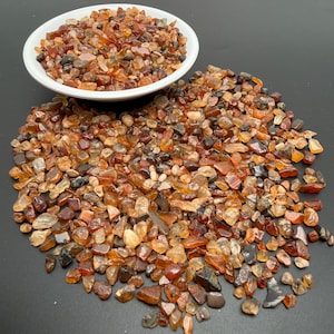 May include: A white ceramic bowl filled with small, polished, tumbled stones in various shades of orange, amber, brown, and translucent yellow. A large pile of the same small, multi-coloured, polished stones is spread out below the bowl.