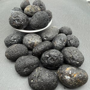 Puede incluir: Una colección de piedras volcánicas oscuras, texturizadas y redondeadas. Algunas piedras están apiladas dentro de un cuenco blanco, mientras que otras están esparcidas por la base. Las rocas presentan una superficie porosa e irregular en tonos negros.