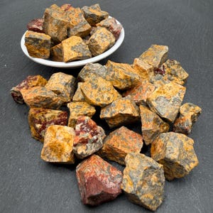 May include: A collection of raw, unpolished stones in shades of mustard yellow, dark grey, and deep red. Some stones are contained within a small white bowl, while others are scattered in a pile, showcasing rough, jagged textures and mineral surfaces.