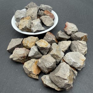 May include: A collection of rough, angular grey stones with earthy brown and orange mineral deposits. Some stones are contained within a small, round white bowl, while others are scattered in a cluster around the base of the bowl.