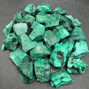 Op de afbeelding: Een verzameling ruwe malachietstenen met levendige groene tinten, natuurlijke gestreepte patronen en gevarieerde texturen. Deze minerale exemplaren vertonen variaties in smaragd- en bosgroen over hun onregelmatige, grillige oppervlakken.