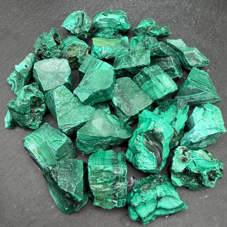 Op de afbeelding: Een verzameling ruwe malachietstenen met levendige groene tinten, natuurlijke gestreepte patronen en gevarieerde texturen. Deze minerale exemplaren vertonen variaties in smaragd- en bosgroen over hun onregelmatige, grillige oppervlakken.
