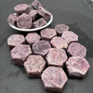 Könnte beinhalten: Eine Sammlung roher, sechseckiger Rubinkristalle in staubigen Rosa- und tiefen Magentatönen mit dunklen Mineraleinschlüssen. Einige Steine sind in einer kleinen weißen Schale gestapelt, während andere auf einer dunklen Oberfläche liegen.