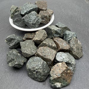 Puede incluir: Una colección de piedras rugosas y angulares en tonos verde oscuro, gris y marrón. Algunas piedras están contenidas dentro de un pequeño cuenco blanco, mientras que otras están esparcidas alrededor de la base, mostrando texturas variadas.