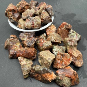 May include: A collection of raw, unpolished stones featuring a mix of earthy green, deep brown, and vibrant orange-red hues. Some stones are contained within a white bowl, while others are scattered around the base, showing rough textures.