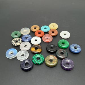 Könnte beinhalten: Eine Sammlung von fünfundzwanzig kreisförmigen Steinperlen mit Mittellöchern in verschiedenen Farben und Texturen, darunter Grün, Blau, Schwarz, Weiß, Braun, Orange und Violett, wobei einige natürliche Mineralmuster und polierte Oberflächen aufweisen.