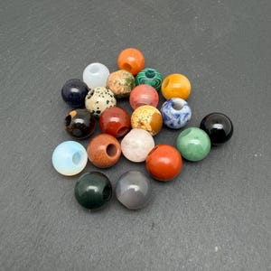May include: A collection of round, drilled beads in various colors and textures, including solid black, white, orange, red, green, and blue, alongside speckled, marbled, and translucent stone-like finishes arranged in a loose cluster.