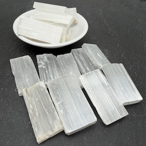 May include: A collection of rectangular selenite crystal slabs with a fibrous, translucent white appearance. Several slabs are stacked inside a white ceramic bowl, while others are arranged on a dark surface, showcasing their natural texture.