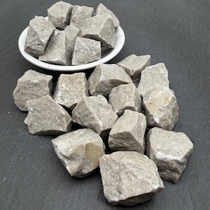 May include: A collection of rough, light grey stones with irregular shapes and textured surfaces. Some stones are contained within a small, round white bowl, while others are scattered in a cluster around the base of the bowl.