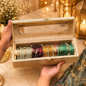 Pode incluir: Uma caixa de joias retangular coberta com tecido bege com uma janela retangular transparente na tampa, contendo várias fileiras de pulseiras circulares finas em várias cores, incluindo verde claro, roxo escuro, dourado e verde esmeralda.