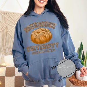 Può includere: Una felpa con cappuccio blu con il testo "SOURDOUGH UNIVERSITY BREADUCATED" e una grafica di una pagnotta di pane a lievitazione naturale.