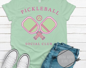 May include: A light green t-shirt with the text "Pickleball Social Club" in pink lettering. The design features two crossed pickleball paddles and a ball in pink and green.