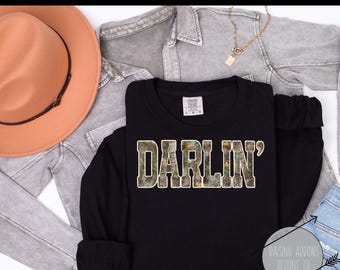 May include: Black long-sleeve shirt with the word "DARLIN'" in a camouflage pattern. The shirt is displayed with a tan hat, a gray denim jacket, and a gold necklace. The shirt is from Raising Arrows Designs Co.
