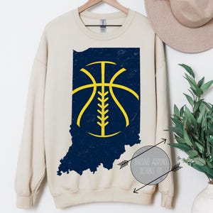 May include: Cream-colored sweatshirt featuring a navy blue Indiana state outline with a yellow basketball design. The design has a distressed, vintage look. The sweatshirt is hanging on a wooden hanger.