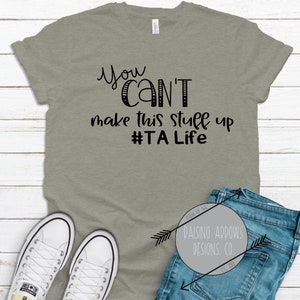 May include: A heather gray t-shirt with black text that reads "You Can't make this stuff up #TALife".