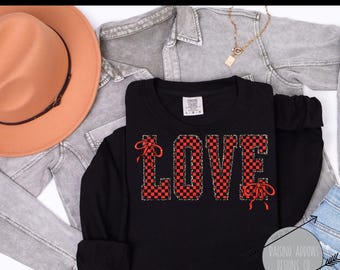 Puede incluir: Camiseta negra de manga larga con la palabra "LOVE" en letras a cuadros rojas y negras. La camiseta tiene lazos rojos y una etiqueta Comfort Colors. Un sombrero color canela y una chaqueta vaquera gris también están en la imagen.