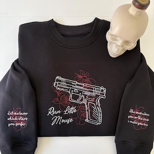 Può includere: Una felpa nera a girocollo con ricami bianchi e rossi di una pistola circondata da rose. Il testo "Run Little Mouse" è ricamato sotto l'arma. Ogni manica presenta ulteriori testi e disegni floreali ricamati.
