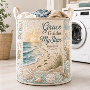 Könnte beinhalten: Ein zylindrischer Wäschekorb aus Stoff mit einer gesteppten Strandszene, die einen Sonnenaufgang, Meereswellen und Fußabdrücke im Sand zeigt. Der Text lautet Grace Guides My Steps Psalm 37:23. Das Design umfasst Muscheln, Seesterne und Strandgras.