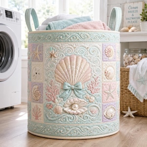 Könnte beinhalten: Ein zylindrischer Wäschekorb aus Stoff mit gestepptem Design und pastellfarbenen Muschel- und Korallenmotiven. Die Außenseite zeigt eine große zentrale Jakobsmuschel mit einer hellblauen Schleife, umgeben von kleineren Muscheln, Seesternen und Wirbelmustern.