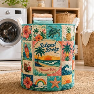 Könnte beinhalten: Ein zylindrischer, gesteppter Wäschekorb mit Patchwork-Design und tropischen Motiven wie Hibiskusblüten, einer Meeresschildkröte, einer Ananas, einem Surfbrett, einer Kokosnuss, einem Regenbogen und einem Seestern. Der mittige Text lautet Island Breeze Tropical Vibes.