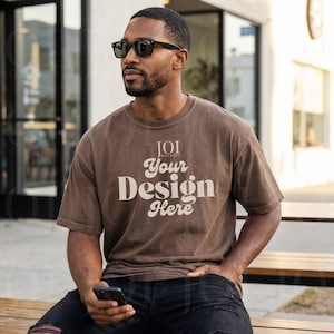 May include: A brown short-sleeved t-shirt featuring the text JOI MOCKUPS Your Design Here in a cream-coloured font. The shirt has a crew neckline and a relaxed fit, worn with black jeans and dark-framed sunglasses in an outdoor setting.
