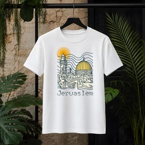 Puede incluir: Una camiseta blanca de manga corta con una ilustración gráfica del horizonte de Jerusalén con un sol y una cúpula amarillos. El diseño utiliza líneas azul oscuro para representar edificios y una torre, con la palabra Jerusalem en fuente punteada debajo.