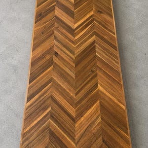 May include: A rectangular wooden tabletop featuring a prominent herringbone pattern. The surface displays natural wood grain variations in shades of medium to dark brown, arranged in a precise V-shaped orientation across the entire flat panel.