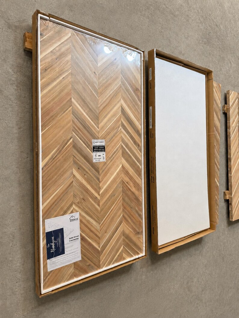 May include: Two rectangular solid wood countertops packaged in cardboard frames. The left countertop features a light brown herringbone pattern with a visible product label reading "Solid Wood Countertop" and "Howiz". The right panel is white.