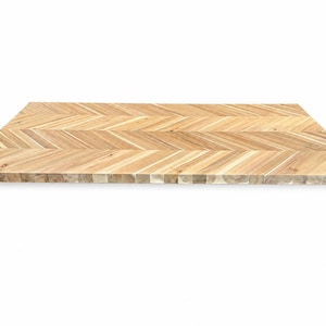 May include: A rectangular wooden board featuring a light brown herringbone pattern. The surface displays natural wood grain variations with alternating angled slats, creating a consistent geometric design across the entire flat top surface.