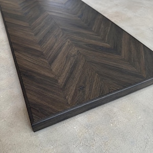 May include: A rectangular wooden board featuring a dark brown finish with a prominent herringbone pattern. The surface displays visible wood grain textures throughout, and the edges are framed with a matching dark wood border.