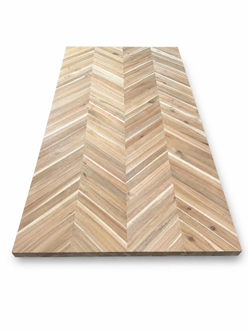 May include: A rectangular wooden tabletop featuring a light brown natural wood grain with a distinct chevron pattern. The surface is composed of multiple planks arranged in a V-shaped design, showcasing varied wood tones and textures.