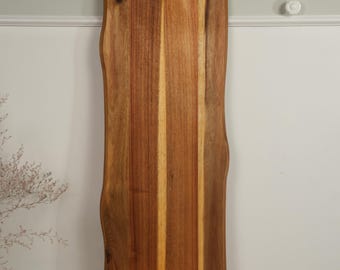 May include: A long, rectangular wooden serving board featuring a natural live edge on both sides. The board is crafted from wood with visible grain patterns in shades of brown and tan. It includes two rectangular cut-out handles at the top and bottom.