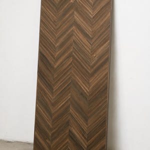May include: A tall, rectangular wooden panel featuring a dark brown finish with a prominent herringbone pattern. The wood grain is visible throughout the surface, and the panel is framed by a thin, matching dark brown border along its edges.