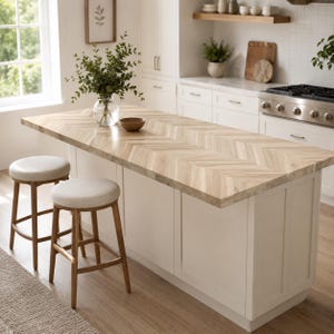 May include: A kitchen island with a light wood countertop featuring a chevron pattern, paired with two wooden stools that have round, beige upholstered seats. A clear glass vase with green eucalyptus branches sits on the countertop.