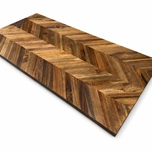 May include: A rectangular wooden board featuring a prominent chevron pattern. The surface displays a mix of light and dark brown wood grain tones, arranged in a V-shaped orientation that runs the length of the board, creating a textured appearance.
