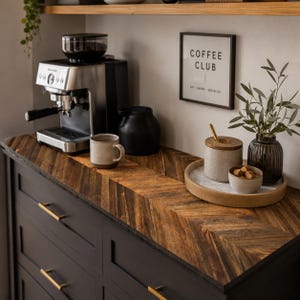 May include: A dark charcoal cabinet with gold handles features a herringbone wood countertop. On top sits a silver espresso machine, a speckled ceramic mug, a black pitcher, and a round wooden tray holding a ceramic jar, a bowl of brown sugar, and a vase.