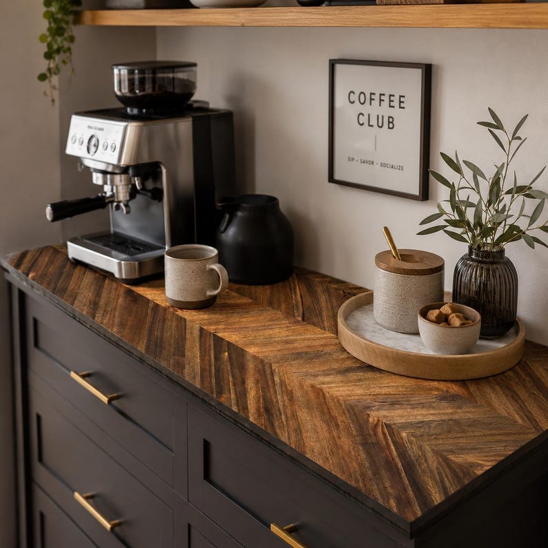 May include: A dark charcoal cabinet with gold handles features a herringbone wood countertop. On top sits a silver espresso machine, a speckled ceramic mug, a black pitcher, and a round wooden tray holding a ceramic jar, a bowl of brown sugar, and a vase.