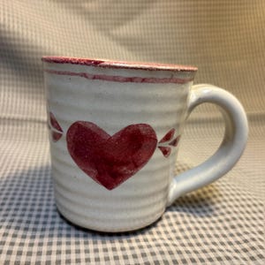 May include: A cream-colored ceramic mug with a ribbed texture, featuring a hand-painted dark red heart on the front and a matching red rim. The mug has a sturdy, curved handle on the right side and a simple, functional design for holding beverages.