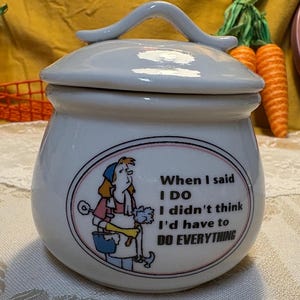 May include: A white ceramic jar with a lid and a handle, featuring a graphic of a person cleaning and the text: When I said I DO I didn't think I'd have to DO EVERYTHING. The jar has a rounded shape and a smooth, glossy finish.